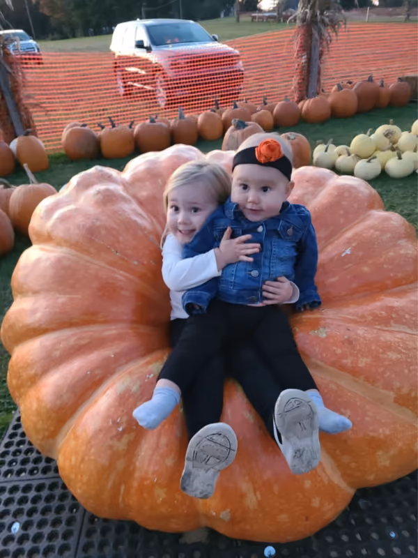 Two young children sitting and hugging on a large orange pumpkin outdoors at a pumpkin patch.