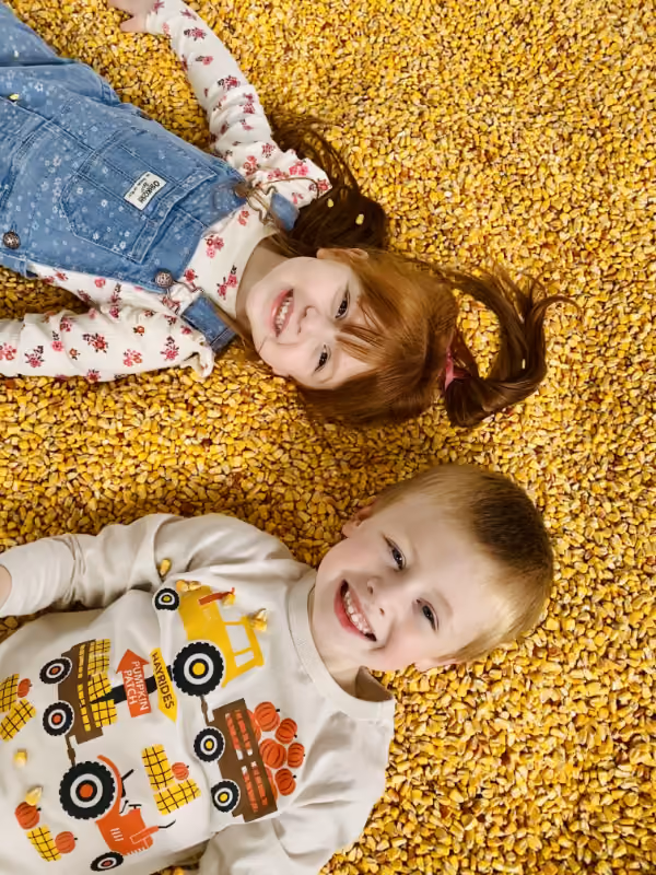 Two young children smiling and lying on a large pile of yellow corn kernels.