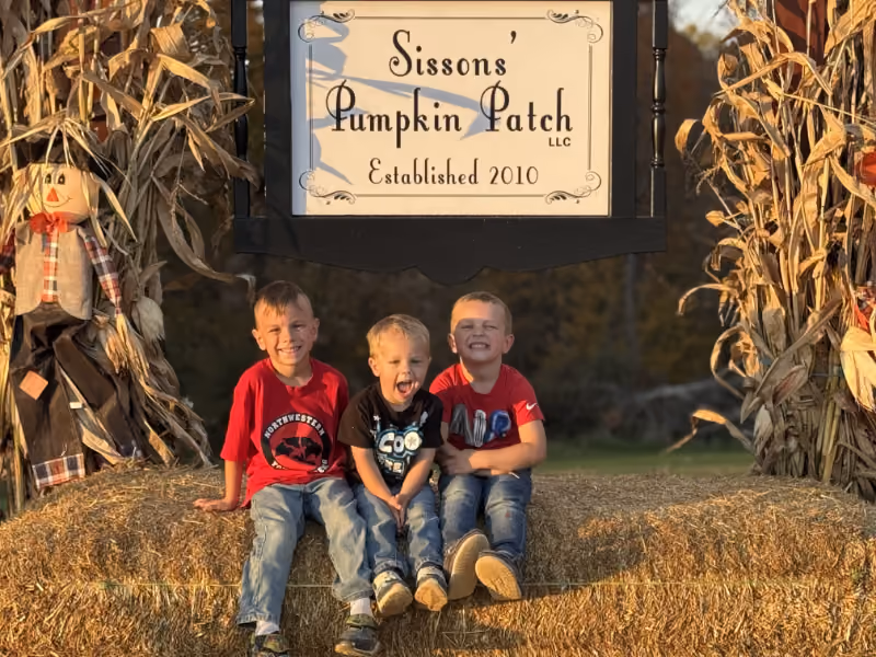 Three boys sitting on hay bales under a sign for Sisson's Pumpkin Patch with scarecrows nearby.