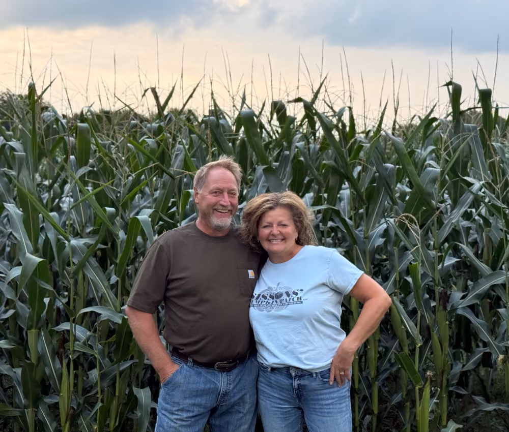Smiling man and woman standing together in front of a tall green cornfield under a cloudy sky.