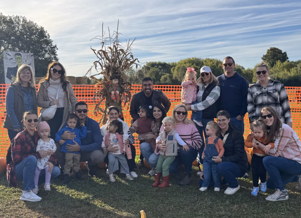 Group of adults and children posing outdoors near a scarecrow and orange safety fence under a blue sky.