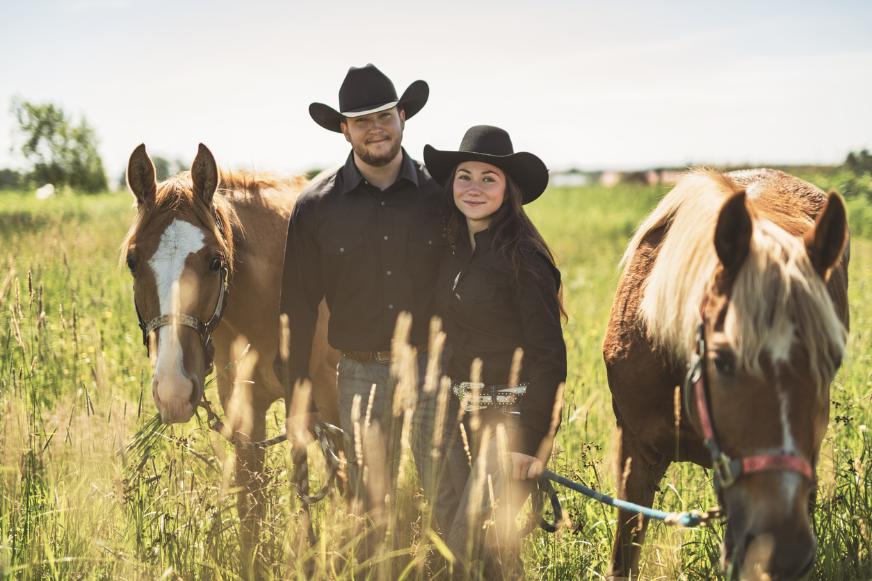 A man and woman in black cowboy hats and shirts standing close together in a grassy field, each holding the reins of a brown horse with white markings on the face, under a clear sky.