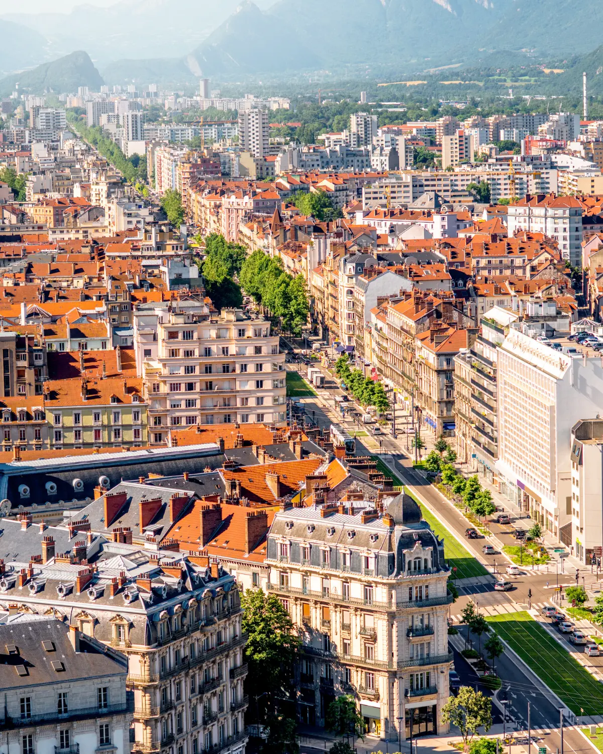 Vue aérienne d'une ville au pied des montagnes, immeubles à toits de tuiles et avenue bordée d'arbres