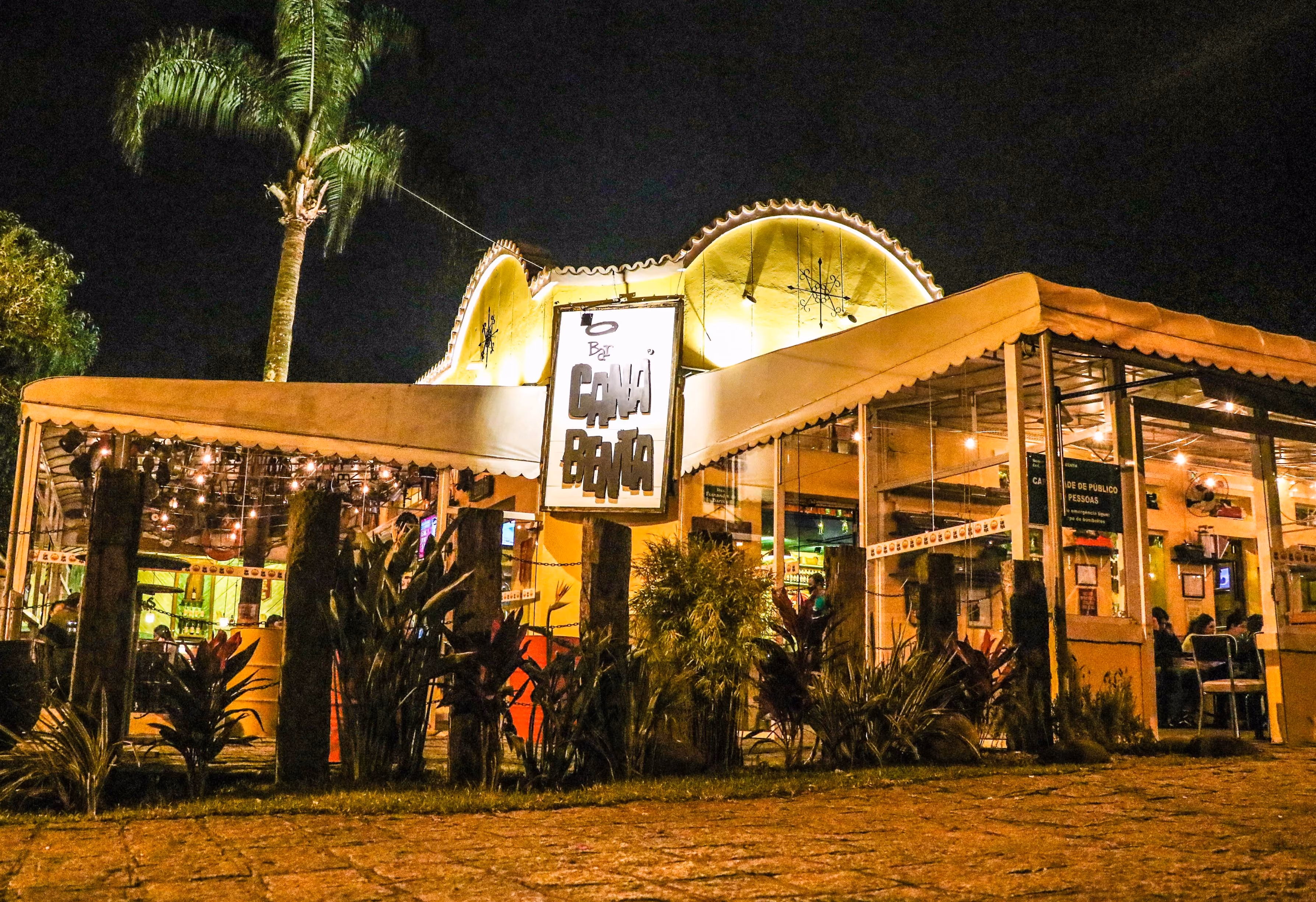 Fachada iluminada à noite de um bar com placa central e plantas em volta do estabelecimento.