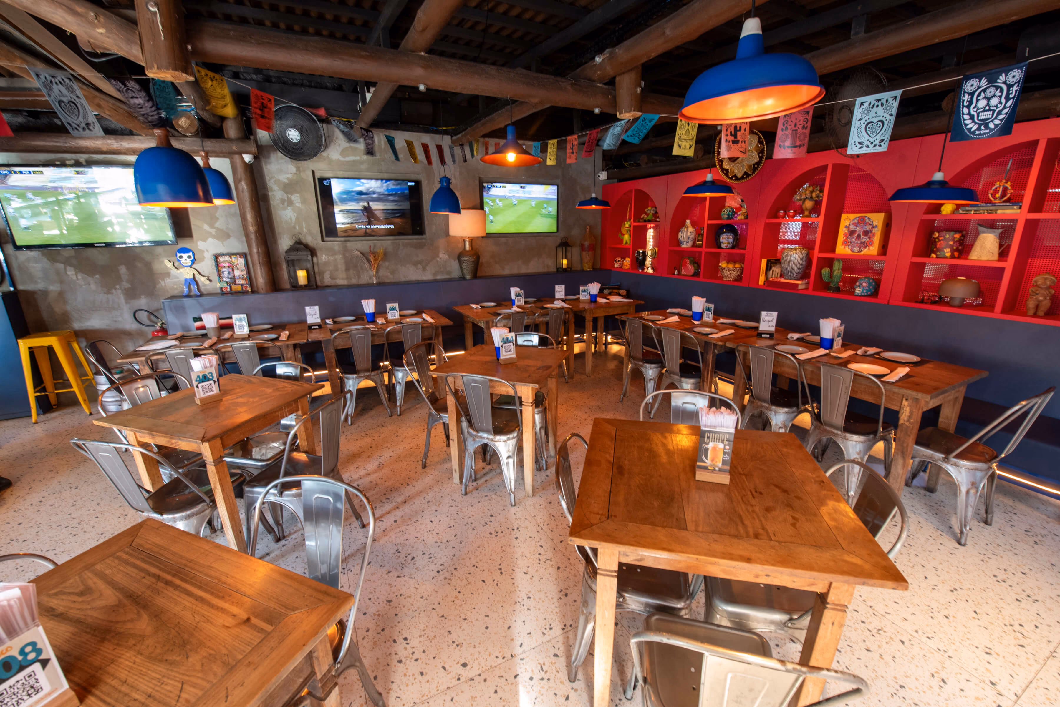 Interior de restaurante com mesas e cadeiras de madeira, luminárias azuis, TV e decoração colorida.