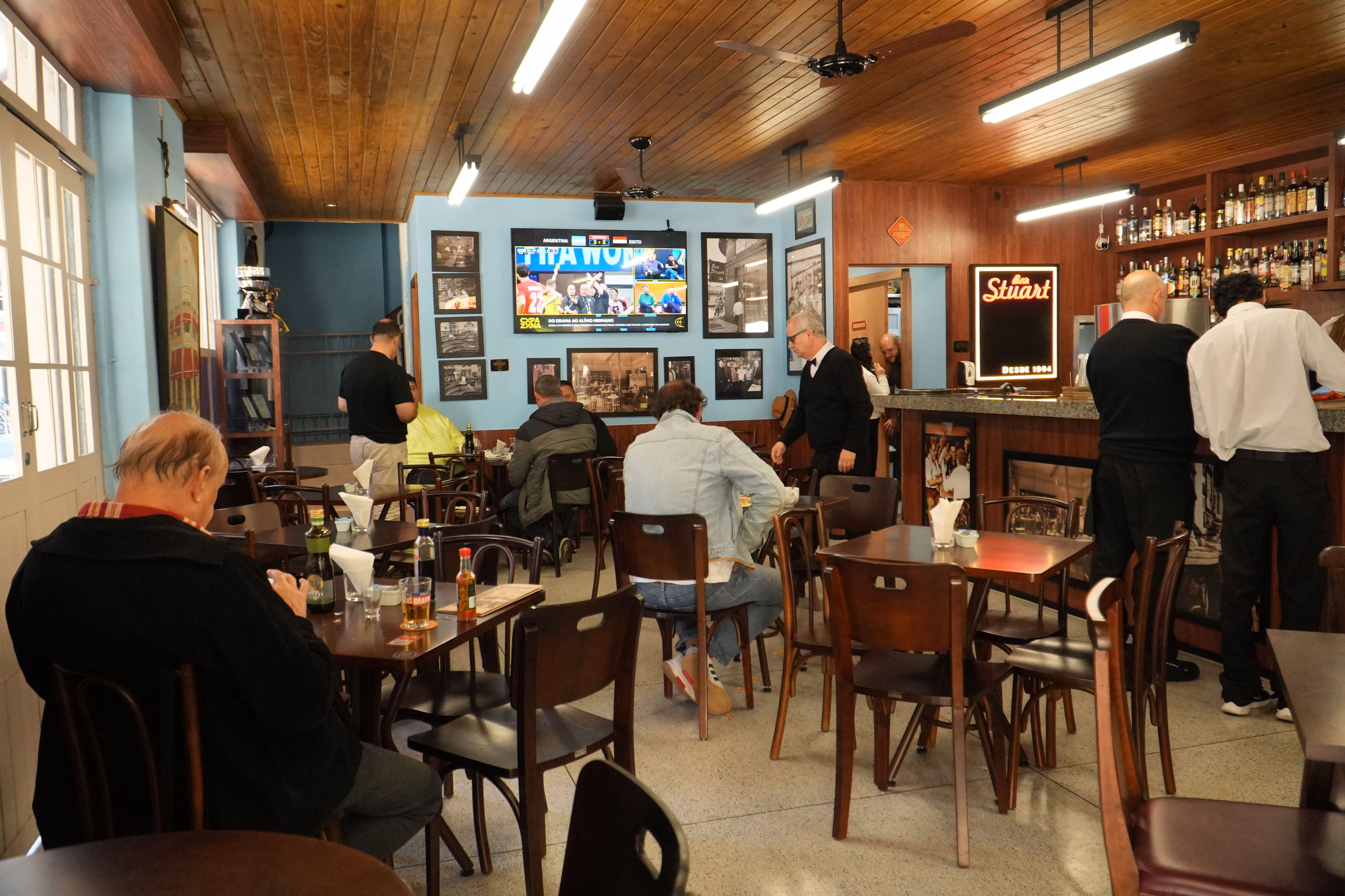 Interior do Bar Stuart com clientes sentados e assistindo a jogo de futebol em TV na parede azul.