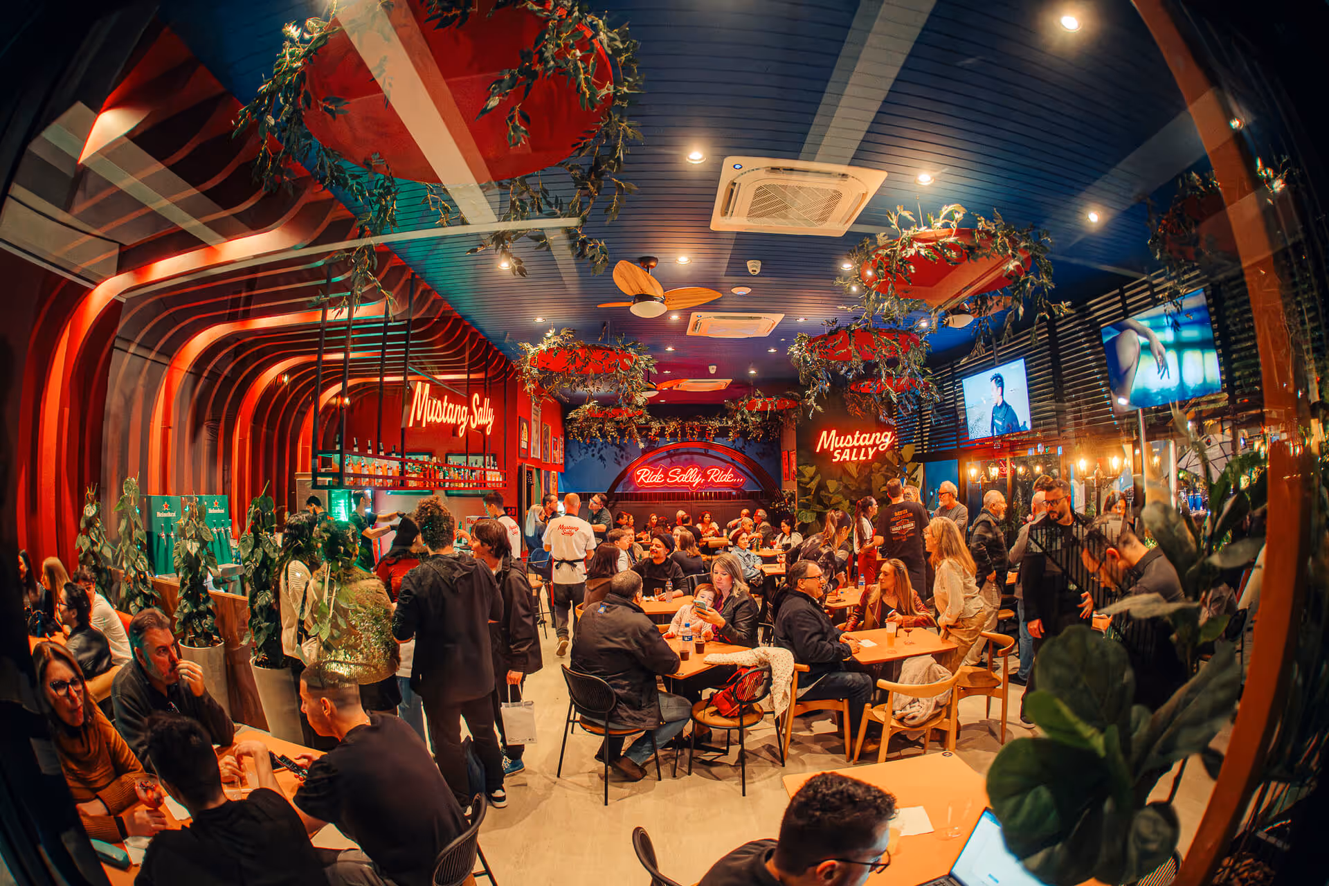 Interior de bar movimentado Mustang Sally com luzes vermelhas, plantas e pessoas socializando em mesas.