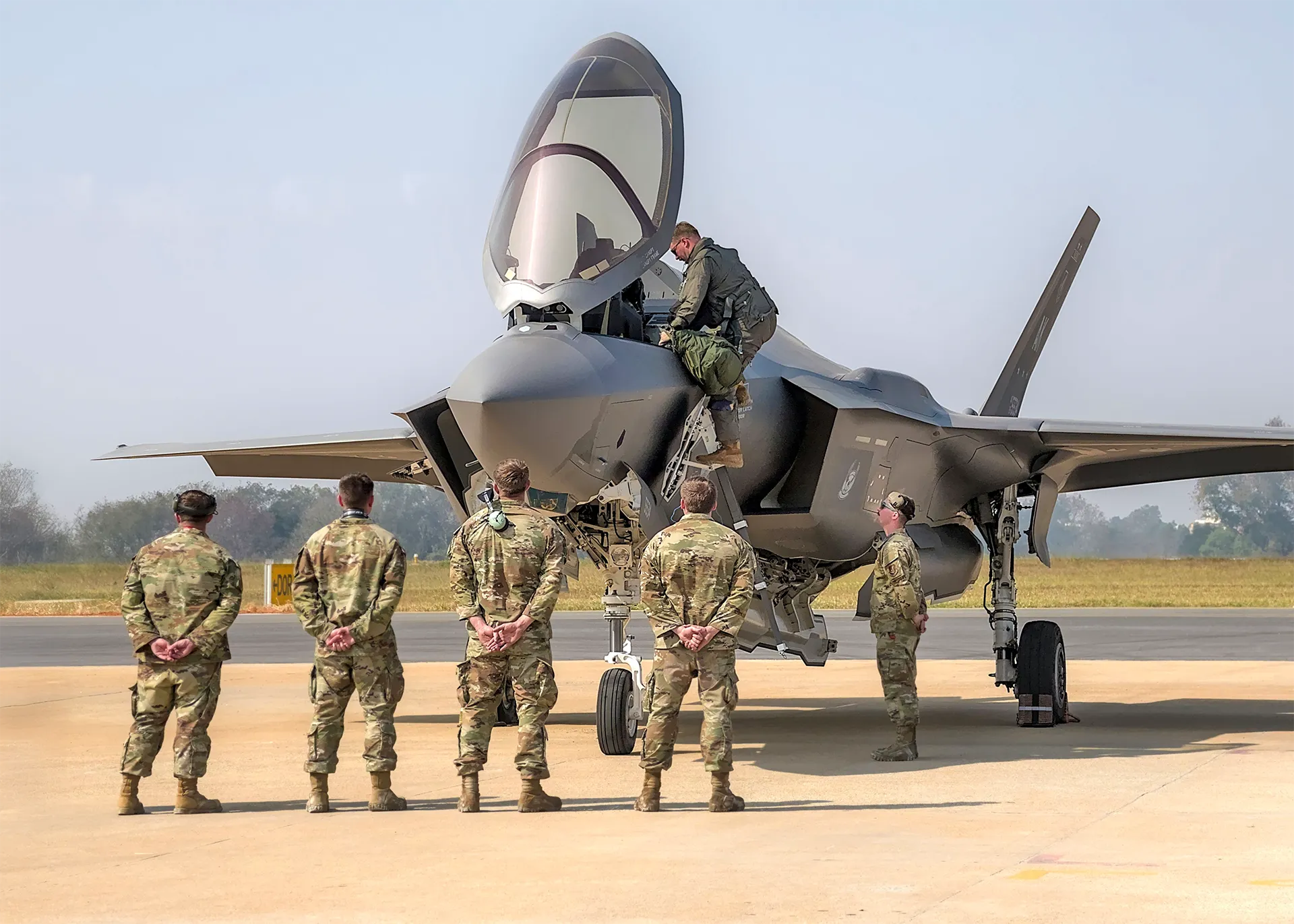 A line of Air Force service members stands at attention on the flight line while a crew member climbs into the cockpit of an F-35 fighter jet.
