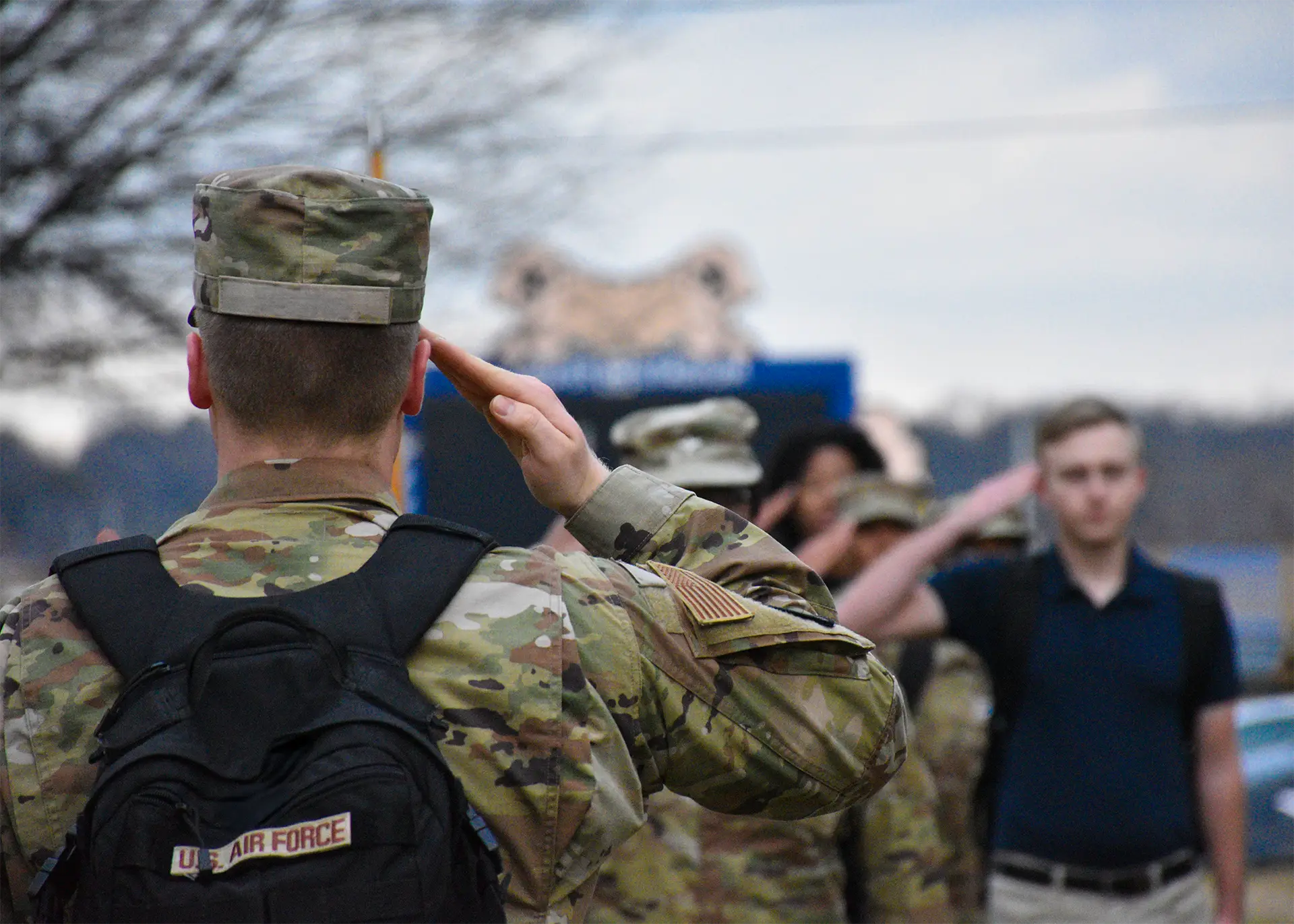 A U.S. Air Force service member is photographed from behind, mid-salute, with a fellow airman visible saluting in the background.