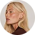 Profile portrait of a blonde woman with earrings wearing a brown top on a light background.