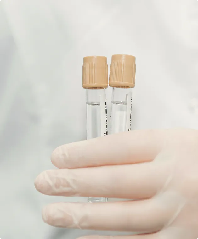 Gloved hand holding two clear test tubes with beige caps against a blurred light background.