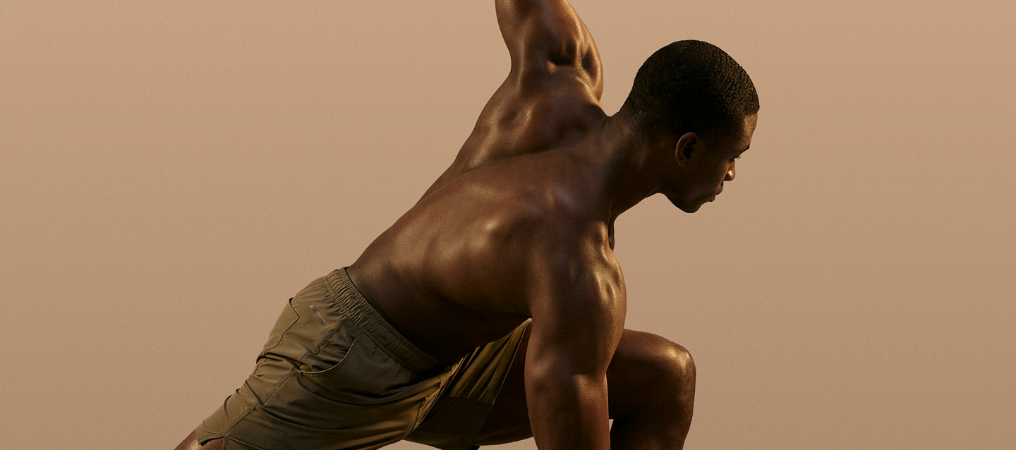 Muscular man in tan shorts poised in a dynamic low lunge against a plain beige background.