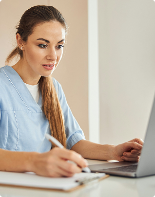 Woman in blue scrubs writing on clipboard while using a laptop at a white desk.