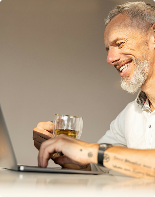 Smiling older man holding a glass of drink and using a laptop at a table.