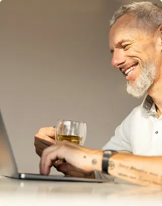 Smiling older man holding a glass of drink and using a laptop at a table.