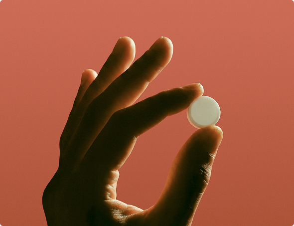 Hand holding a small round white pill against a reddish background.