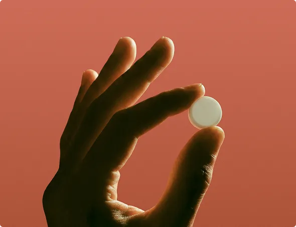 Hand holding a small round white pill against a reddish background.