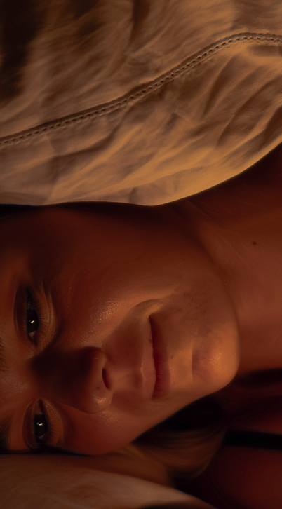 Close-up of a woman's face resting sideways with soft lighting and beige fabric above her.
