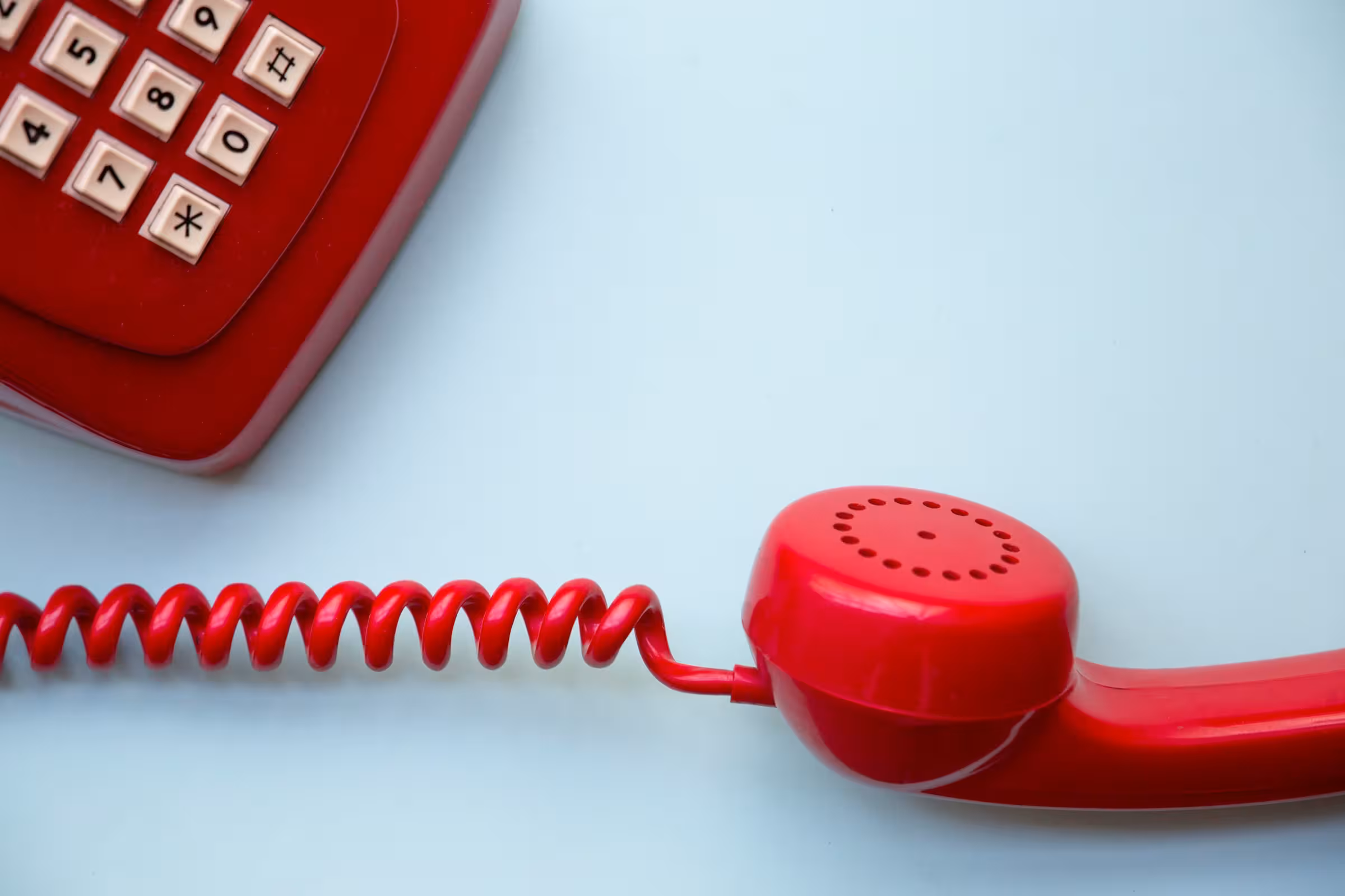Close-up of a red vintage landline telephone handset and coiled cord on a light blue surface, with part of the keypad visible.