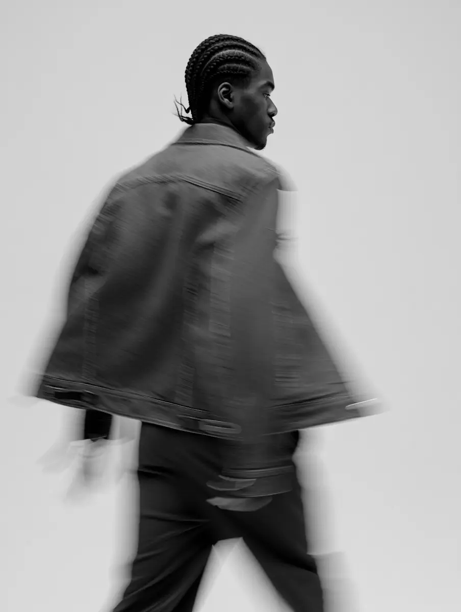 Black and white motion blur photo of a young man with braided hair walking, wearing a denim jacket and dark pants, facing sideways.