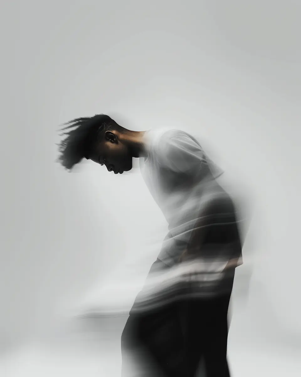 Blurred artistic portrait of a young person with dark skin and textured hair, bending forward against a light gray background with motion blur effect.