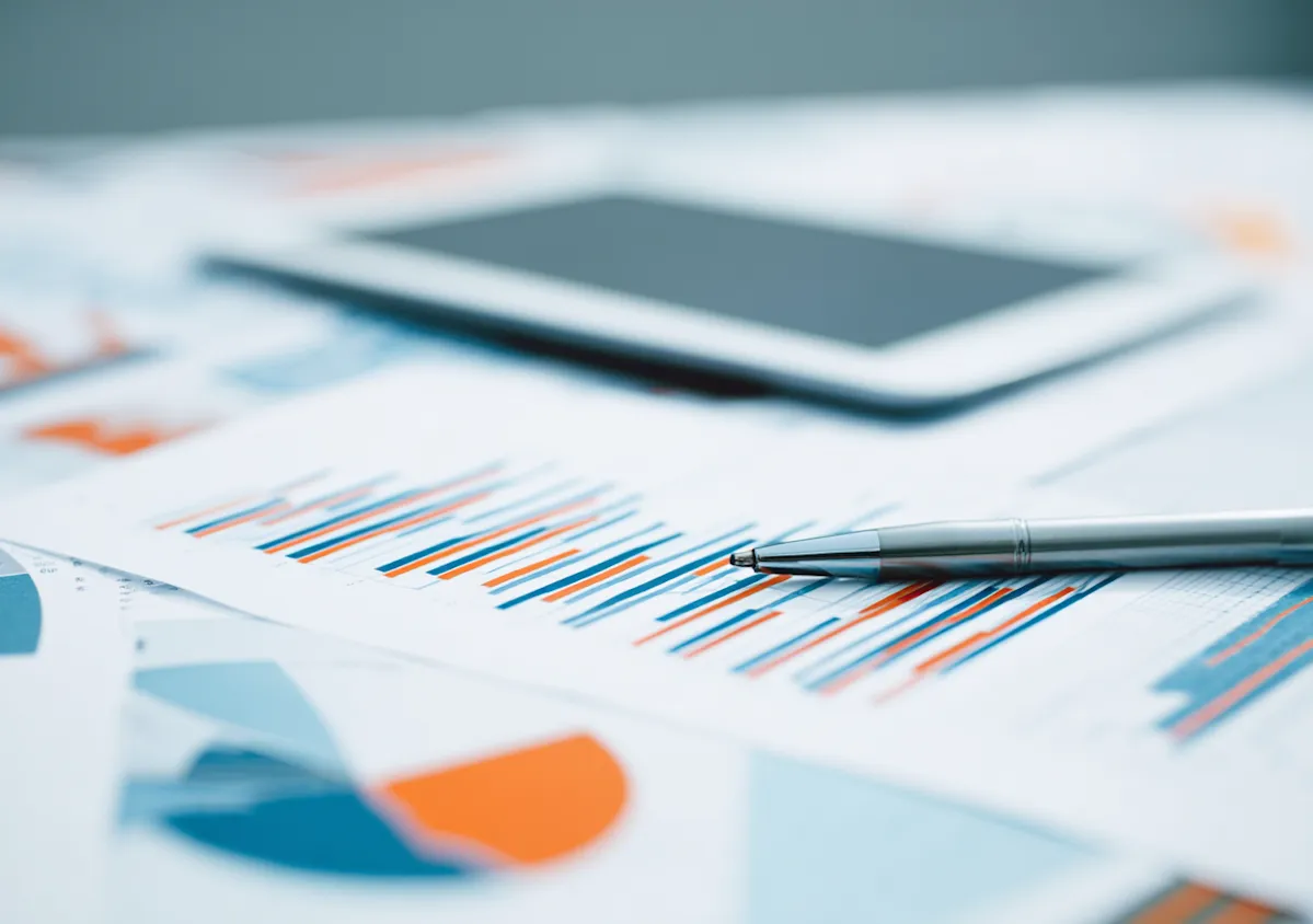 A close-up photograph of a clean office desk surface, featuring a tablet resting on top of several open financial documents and charts.