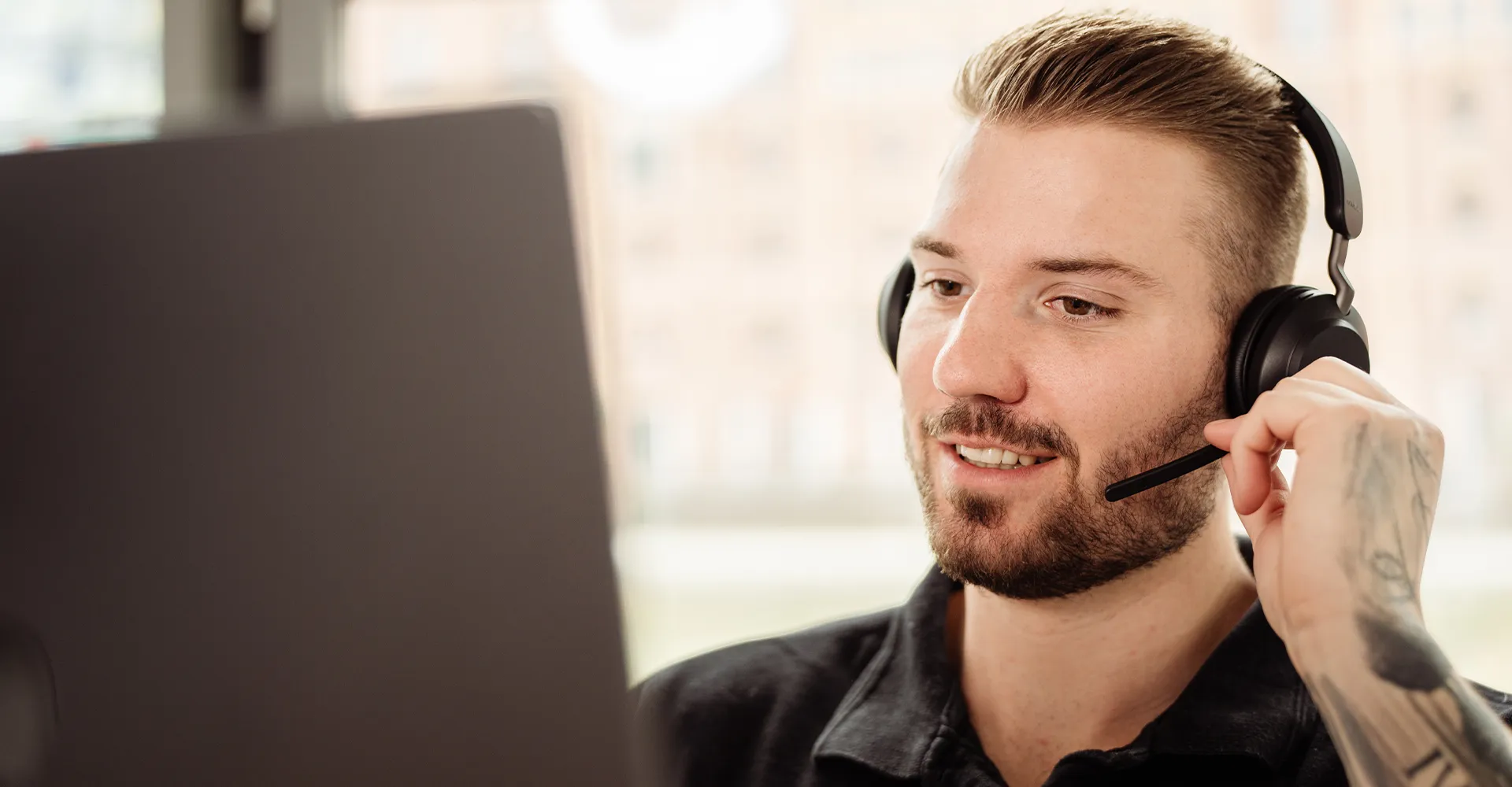 Junger Mann mit Headset bedient Laptop und spricht, tätowierter Arm sichtbar.