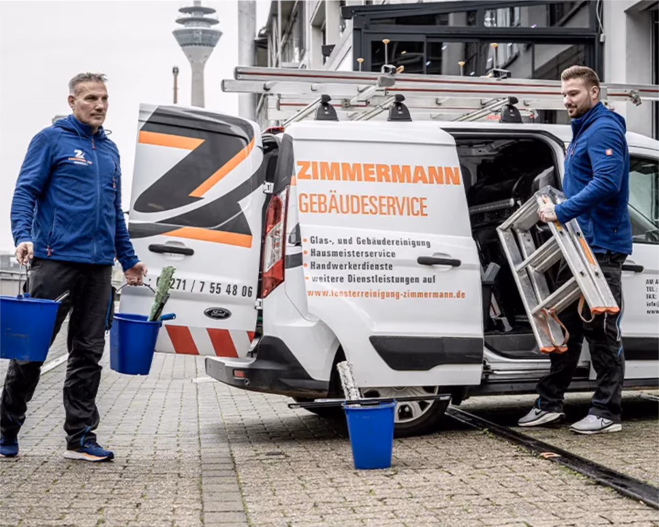 Zwei Männer in blauen Jacken laden Putzutensilien und eine Leiter aus einem Zimmermann Gebäudeservice Fahrzeug aus.