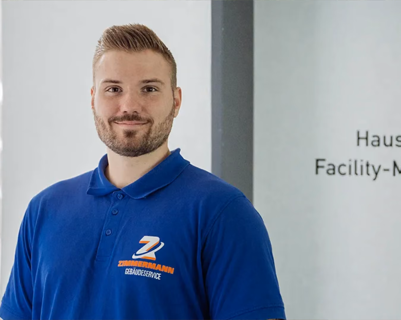 Mann mit blauem Poloshirt mit Zimmermann Gebäudeservice Logo vor weißer Wand.