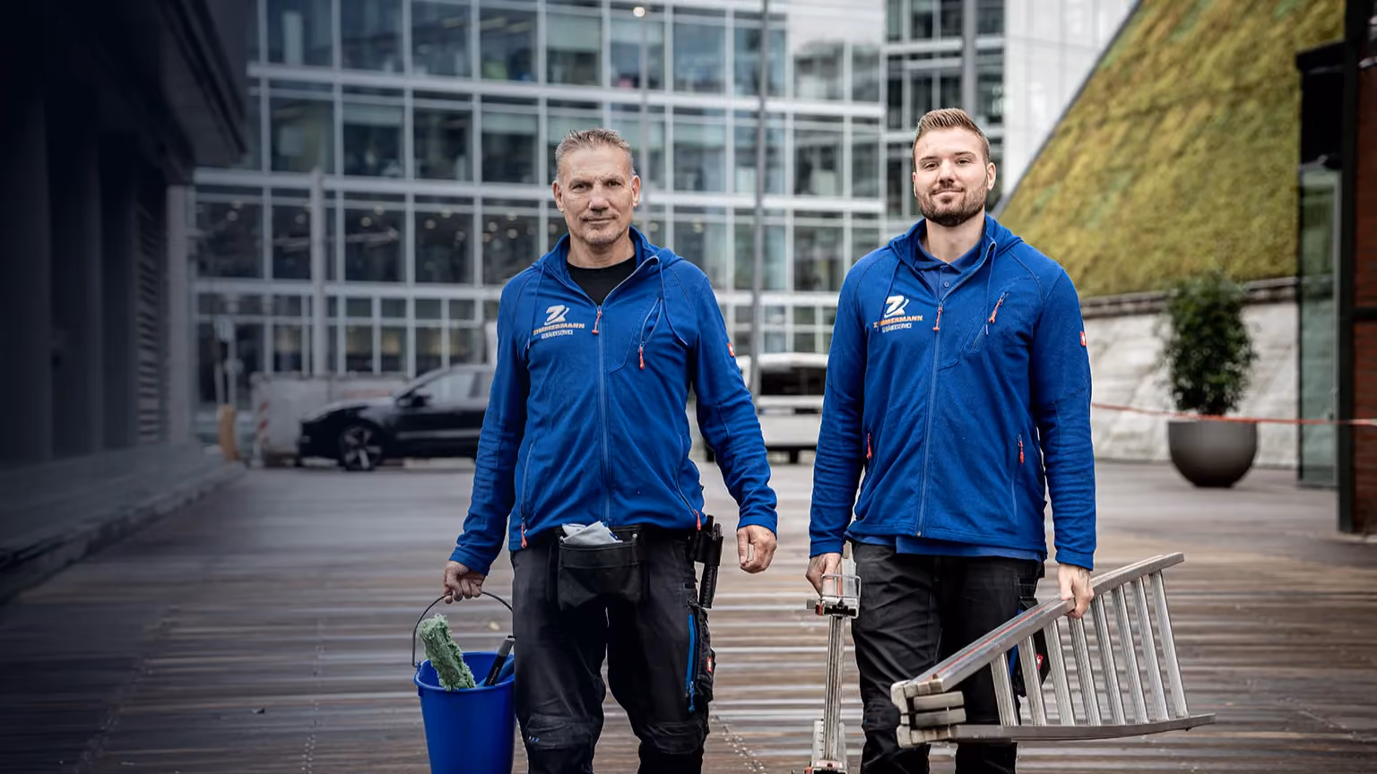 Zwei Männer in blauen Zimmermann-Gebäudeservice-Jacken mit Putzeimer und Leiter vor modernem Gebäude.