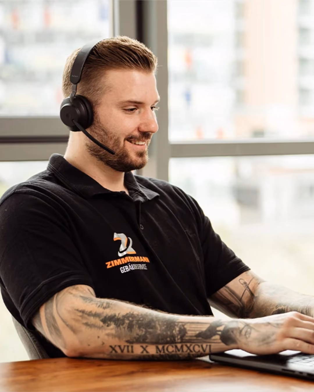 Lächelnder Mann mit Headset und tätowierten Armen tippt auf einem Laptop vor Fenster.
