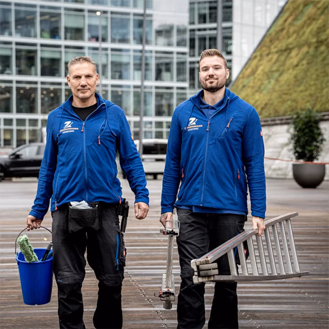 Zwei Männer in blauen Jacken tragen Putzeimer und zusammengefaltete Leiter vor einem Gebäudekomplex.