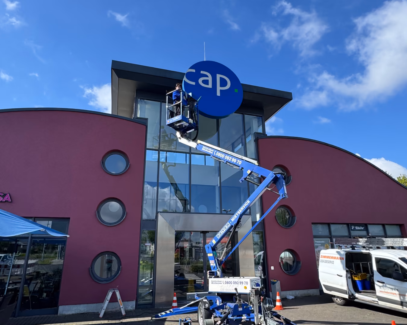 Arbeiter auf Hebebühne reinigt ein rundes CAP-Schild an Gebäudefassade unter blauem Himmel.
