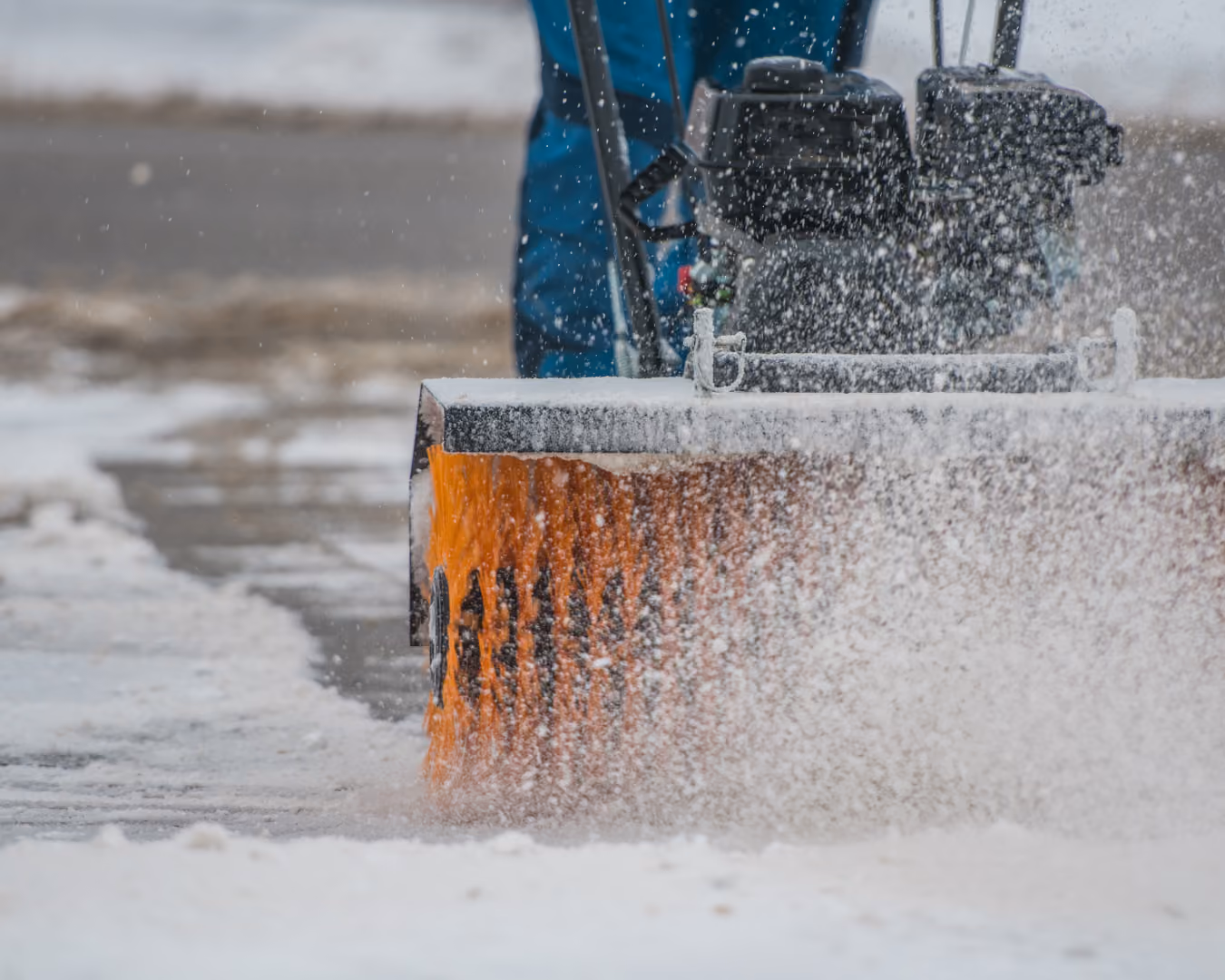 Person using a snow plow machine with an orange brush to clear snow from a sidewalk.