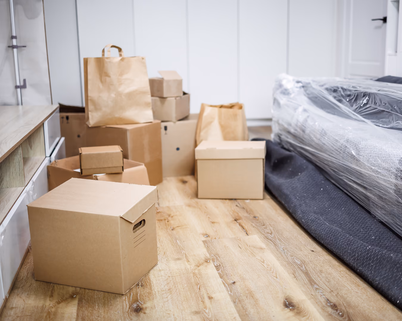 Mehrere Umzugskartons und Papiertüten stehen auf einem Holzboden neben einer verpackten Couch.