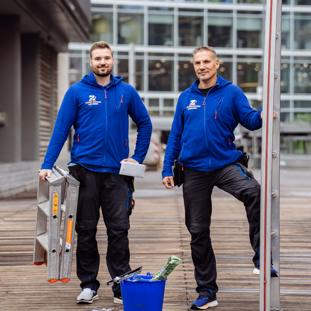 Zwei Handwerker in blauen Jacken stehen mit Reinigungsgeräten auf Holzterrasse vor Gebäude.