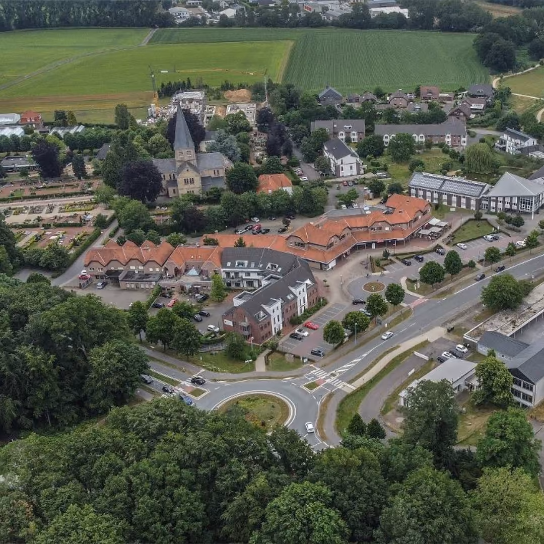 Luftaufnahme einer kleinen Stadt mit einem rundlichen Kreisverkehr im Vordergrund, mehreren roten und weißen Wohngebäuden, einer Kirche mit spitzem Turm, Bäumen und grünen Feldern im Hintergrund.