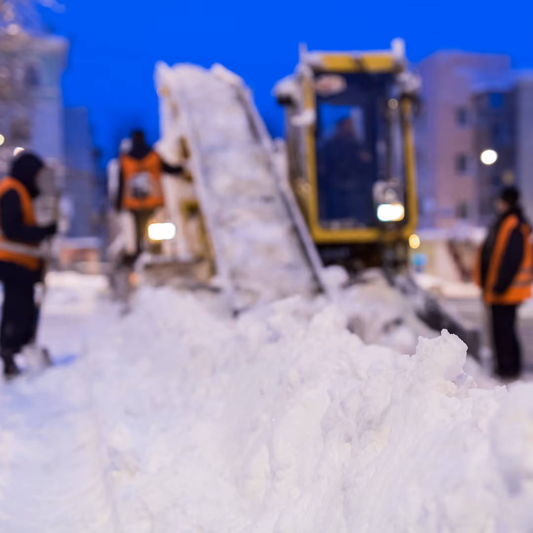 Winterdienst-Maschine hebt Schnee auf und transportiert ihn über ein Förderband, während Mitarbeiter in orangenen Warnwesten neben der Maschine stehen, bei Dämmerung in einer schneebedeckten Stadtumgebung.