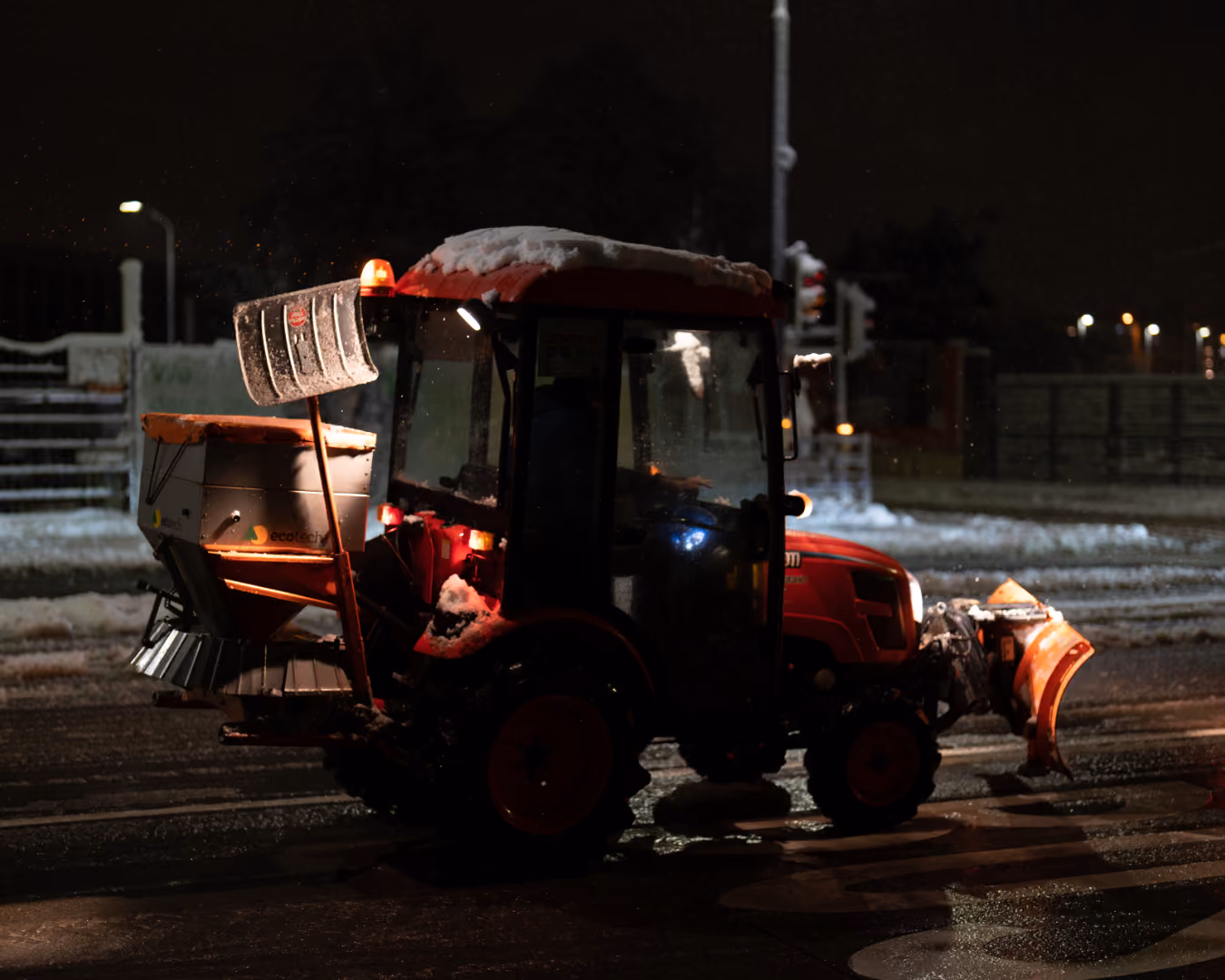 Roter Winterdienst-Traktor mit Schneepflug vorne und Streuapparat hinten bei Nacht auf schneebedeckter Straße.