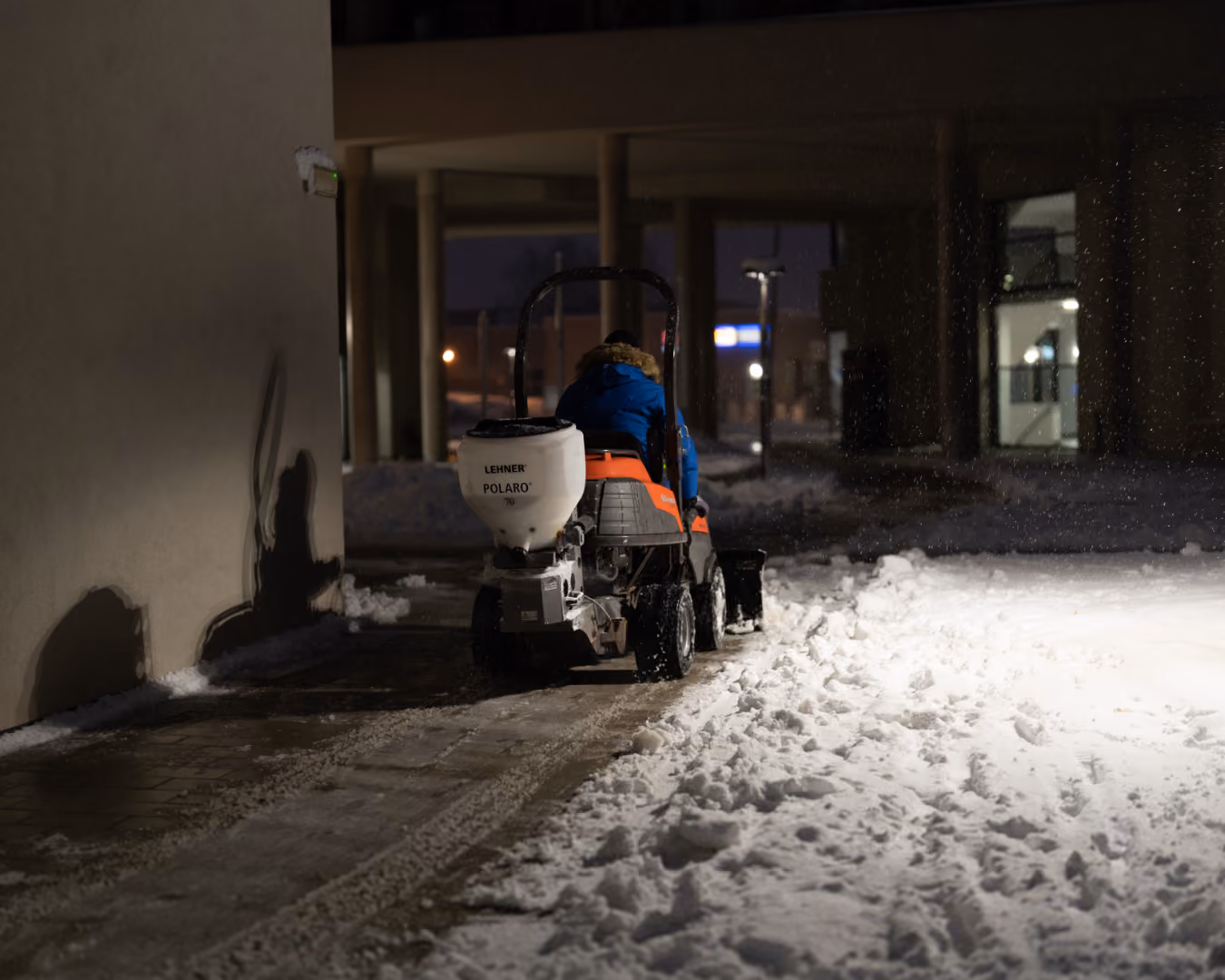 Person im Winter bei Nacht auf einem kleinen Traktor mit Schneeschaufel und Streugutbehälter, räumt und streut eine schneebedeckte Fläche neben einem Gebäude.