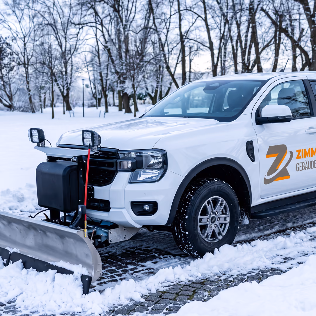 Weißer Geländewagen mit vorne montiertem Schneeräumschild fährt auf gepflastertem Weg zwischen Schnee bedeckten Bäumen.