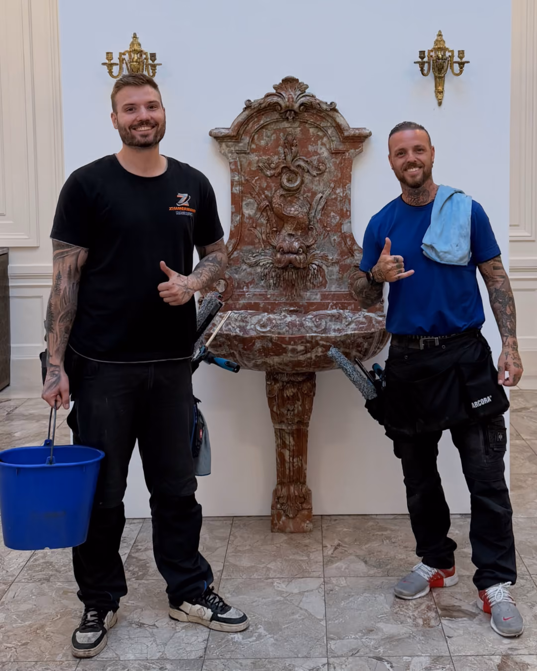 Zwei lächelnde Männer mit Tattoos posieren vor einem antiken, rotem Steinbrunnen mit verzierter Rückwand, sie halten Reinigungswerkzeuge.