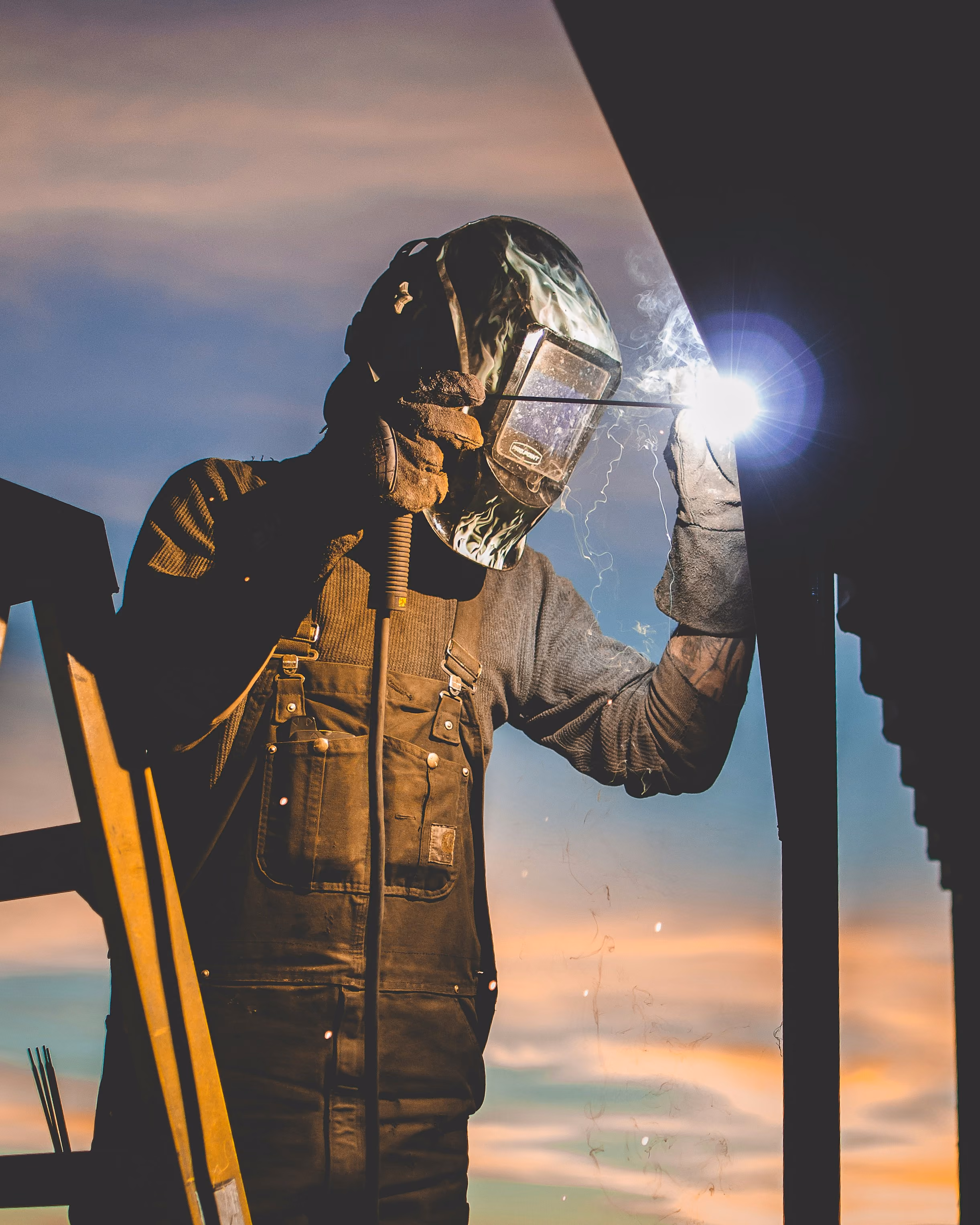 Welder in protective gear working on metal with bright welding light at sunset.