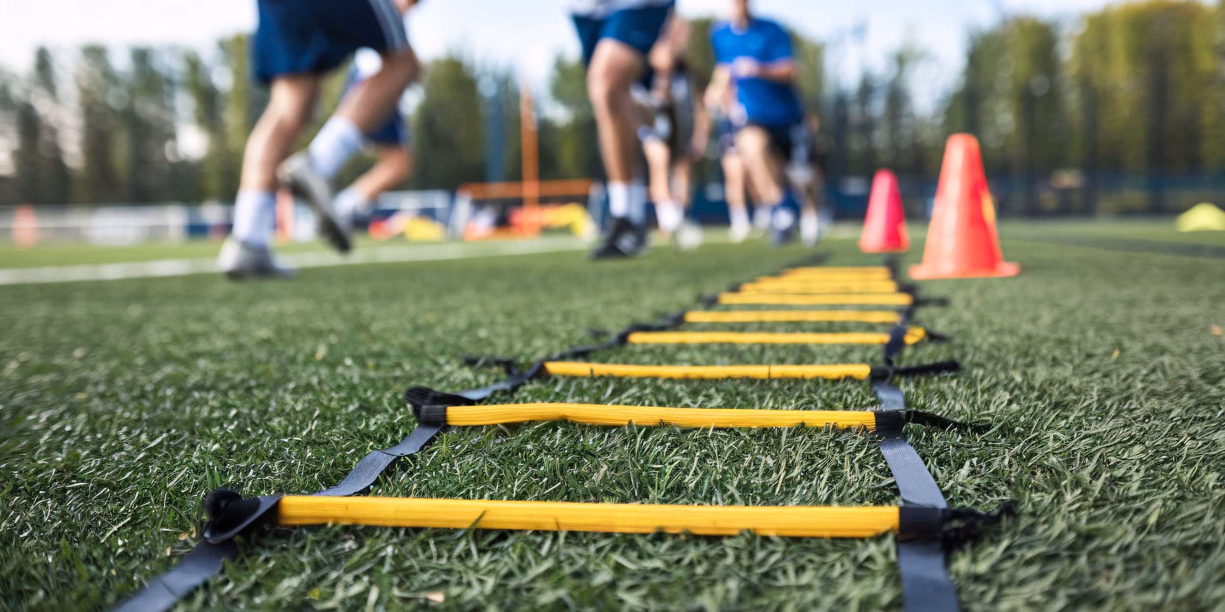 An agility ladder lies on artificial turf with orange cones in the background as people run through a training drill.