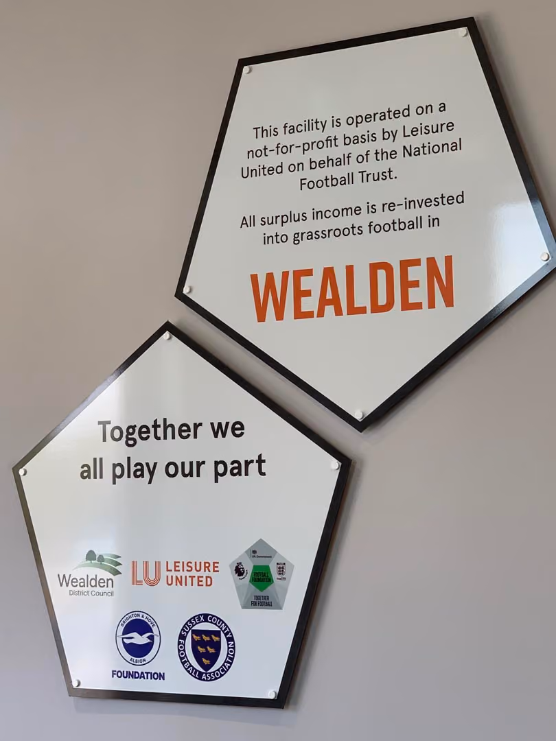 Two shield-shaped signs displaying information about Wealden football facility operated on not-for-profit basis by Leisure United.