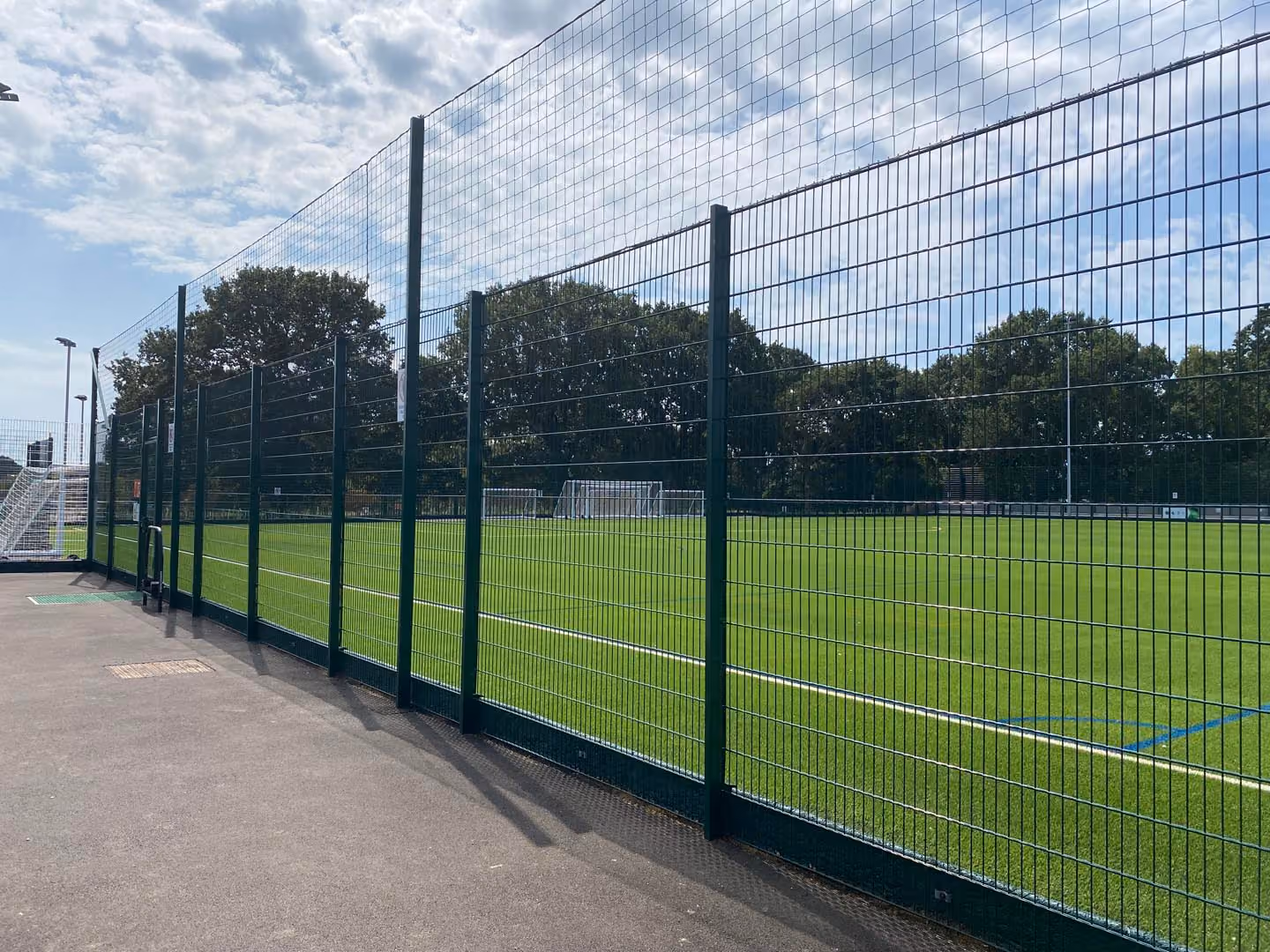 Modern sports facility with green mesh fencing, goal posts, and grass field under blue sky