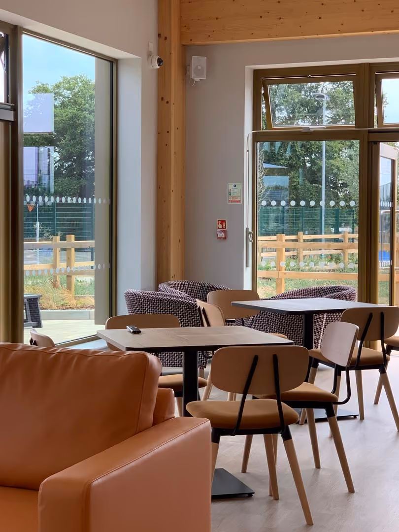 Modern office lounge with brown leather furniture, wooden chairs, tables, and large windows overlooking garden.