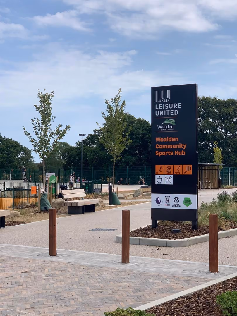 Wealden Community Sports Hub signage at Leisure United sports facility entrance with trees and playground.