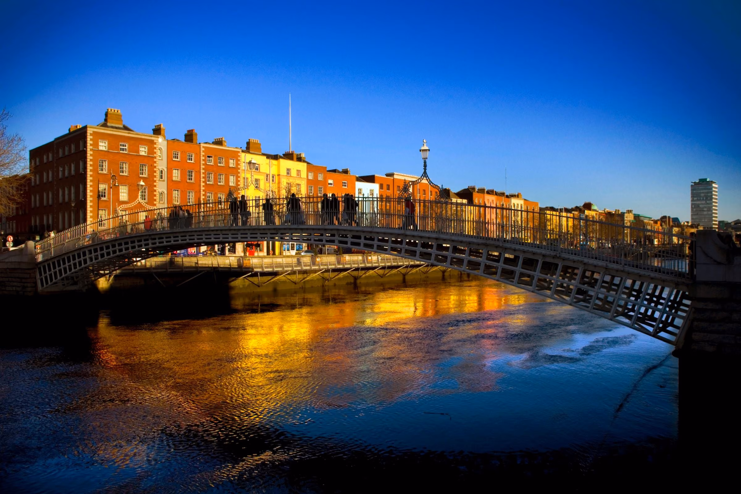 Arched pedestrian bridge over river with colorful buildings and clear blue sky in the background.