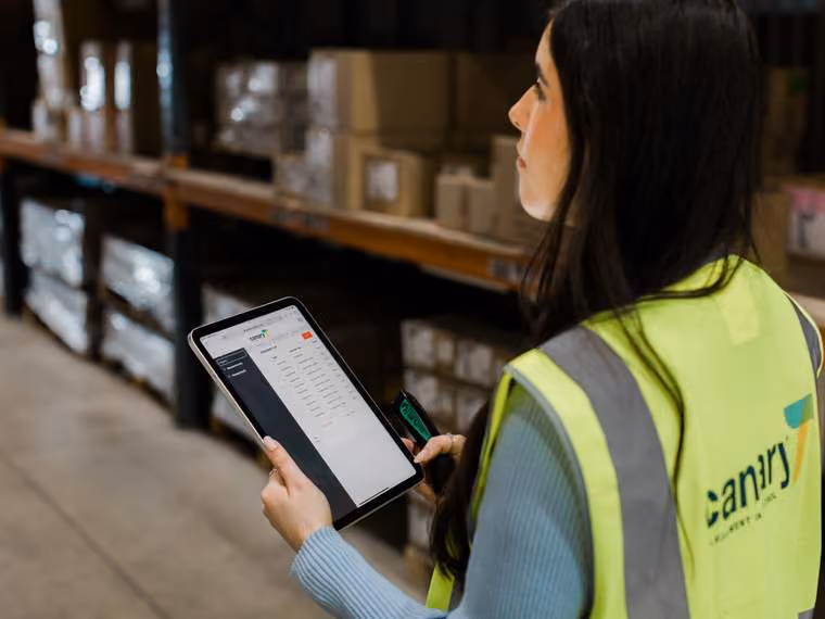 Woman in a high-visibility vest holding a tablet and scanner in a warehouse aisle with stacked boxes.